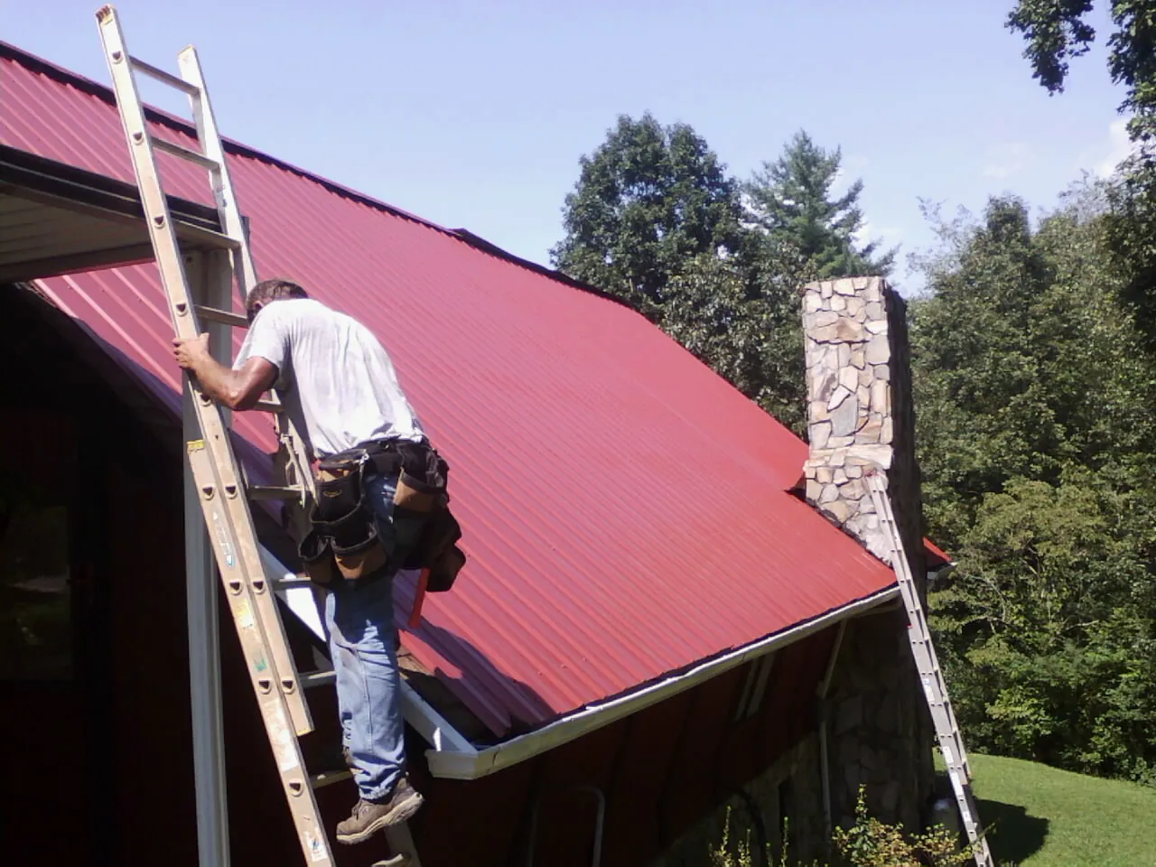 Roofer on a ladder beside a red metal roof and stone chimney — Soffit & Fascia Repair by Roofing Brothers in Dowagiac